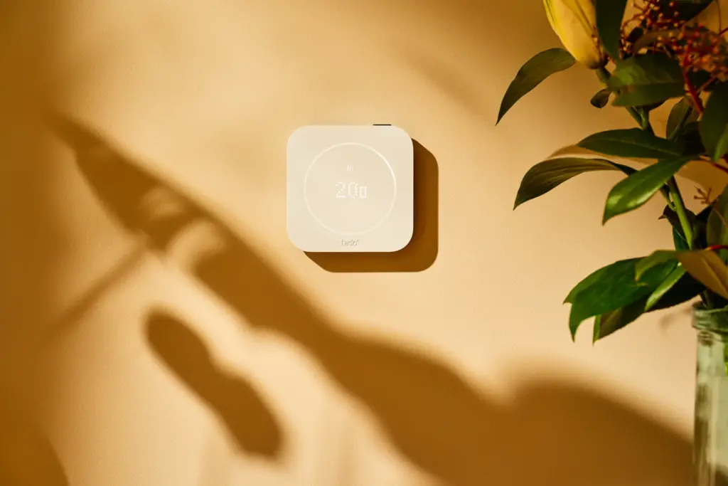 tado Smartes Thermostat X der 2. Generation: mattweißes Wandthermostat mit Lichtring und Temperaturanzeige an einer warm beleuchteten Wand neben einer Pflanze